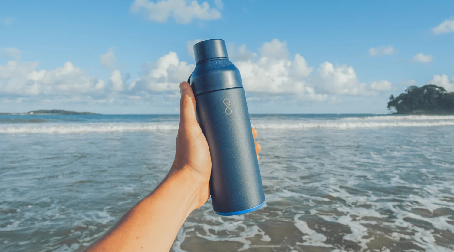 A dark blue Ocean Bottle with a light blue base is held in a person's hand against a backdrop of the ocean with waves and a partly cloudy sky.
