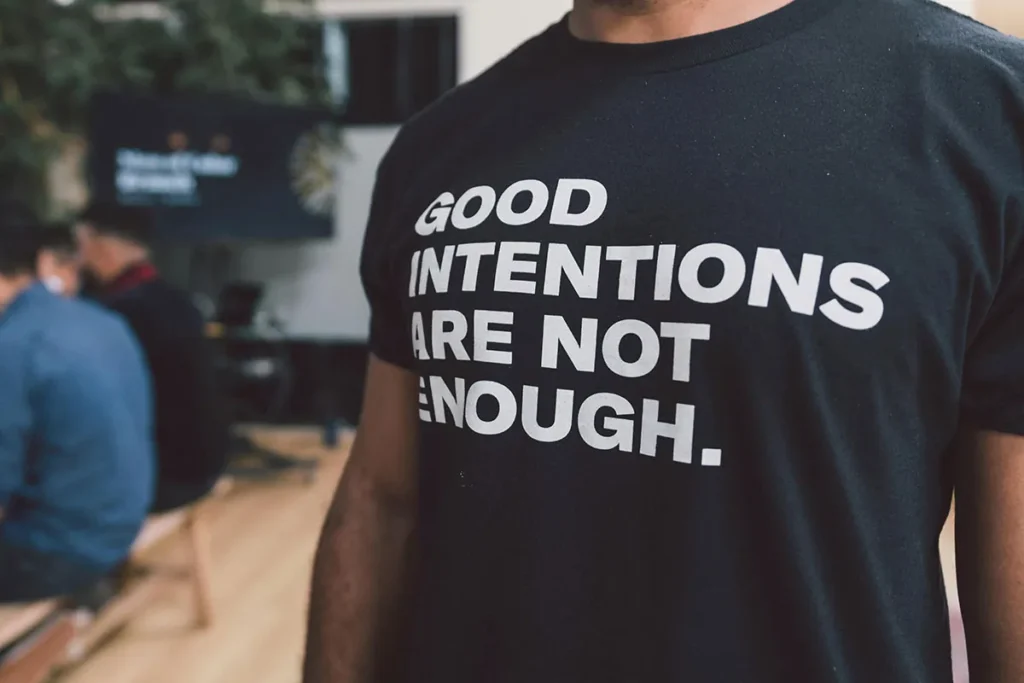 A close-up shot shows a person wearing a black t-shirt with the white text: "GOOD INTENTIONS ARE NOT ENOUGH."